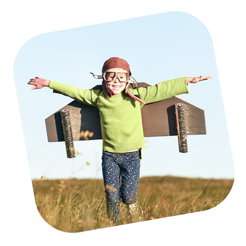 imagem de um garotinho com óculo de aviador e com uma asa de papelão grudada nas costas brincando feliz em área aberta na natureza